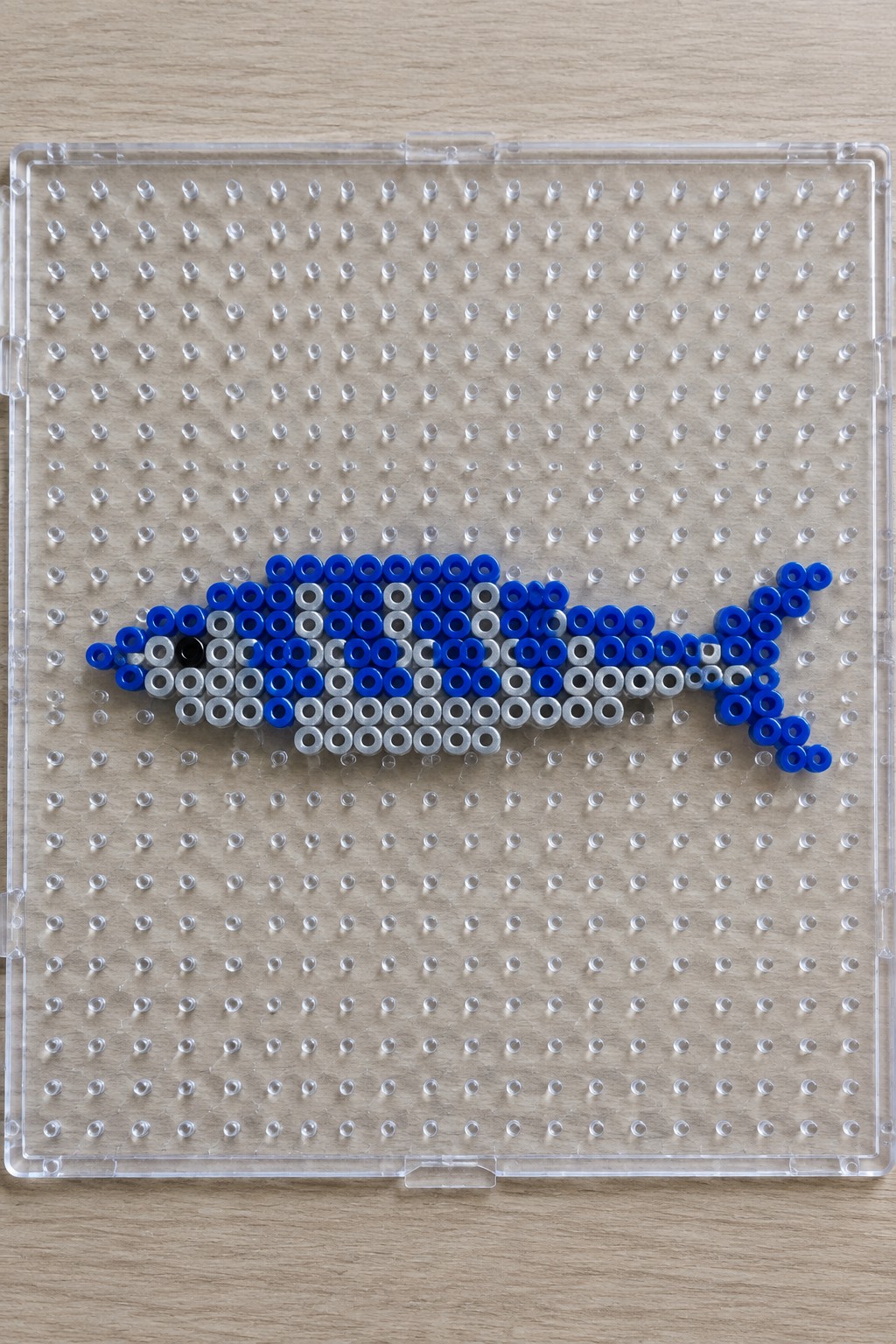 Blue and silver beads forming fish on clear pegboard over wood