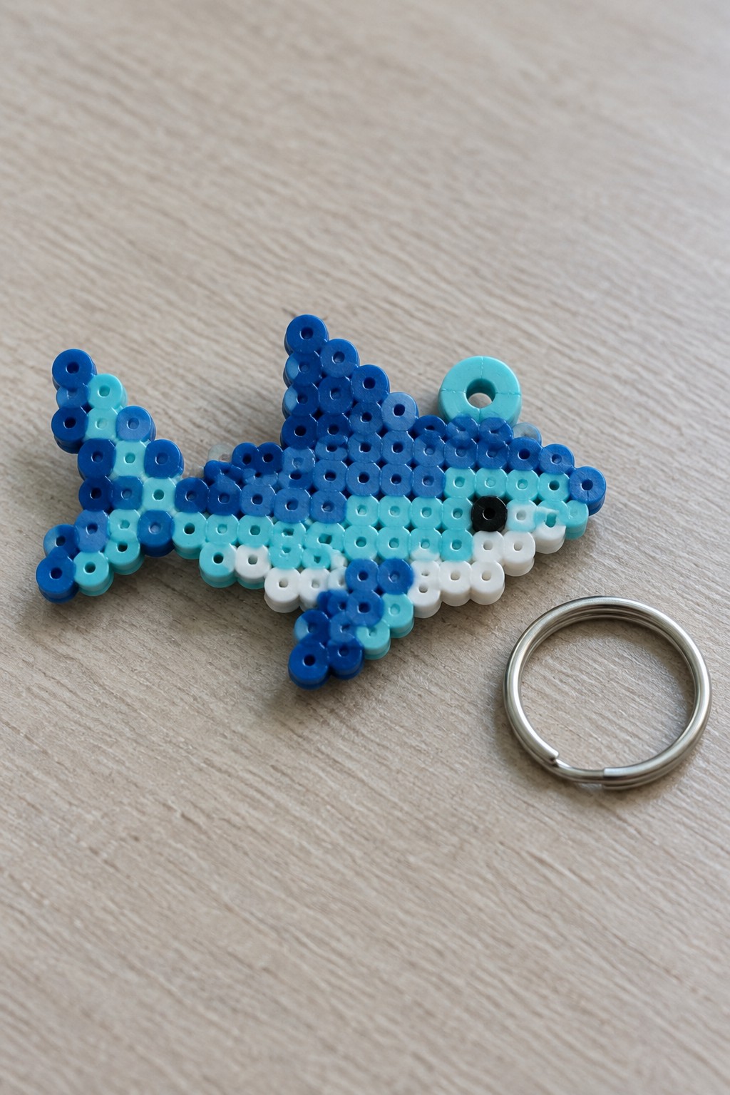 Blue and white perler bead fish keychain beside metal ring on wood surface.