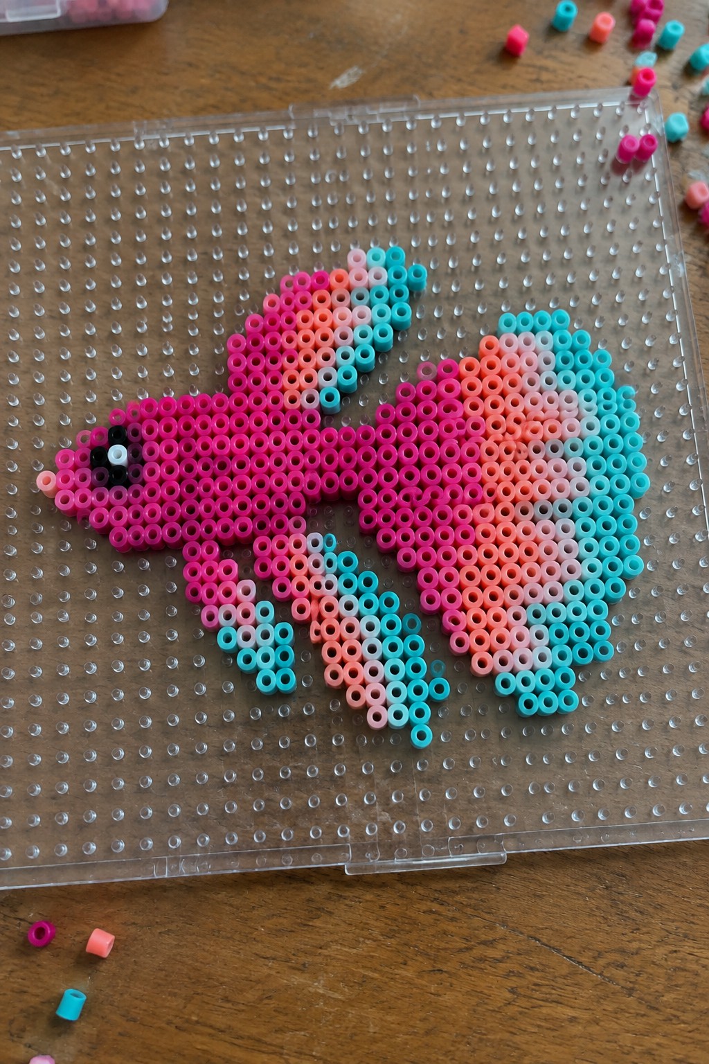 Pink orange cyan perler beads forming butterfly on clear pegboard with loose beads