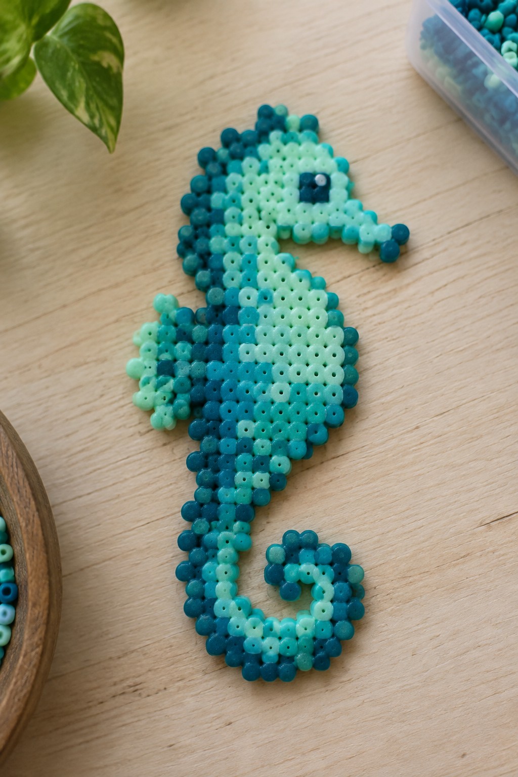 Blue-green perler bead seahorse on wooden table with bead supplies nearby