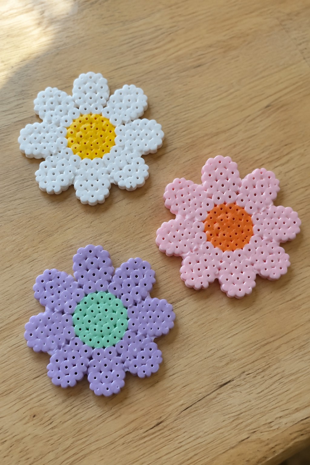 Three Perler bead flowers with white, pink, and purple petals on wood.