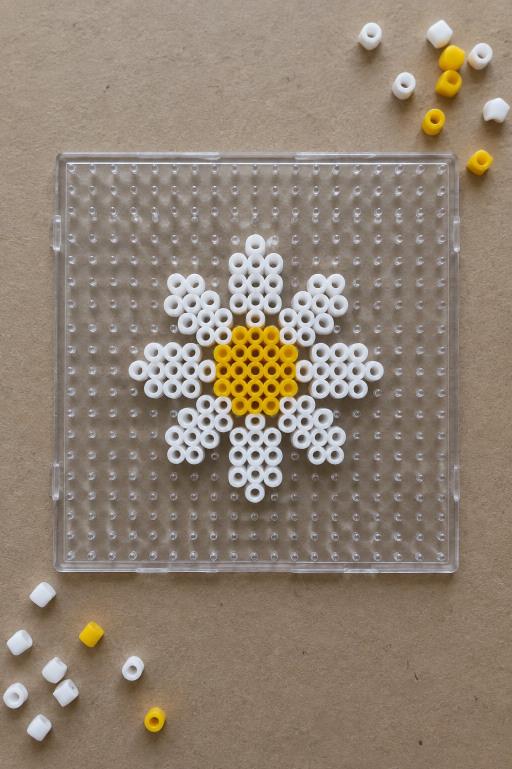 Daisy of white and yellow beads on clear pegboard with loose beads nearby.