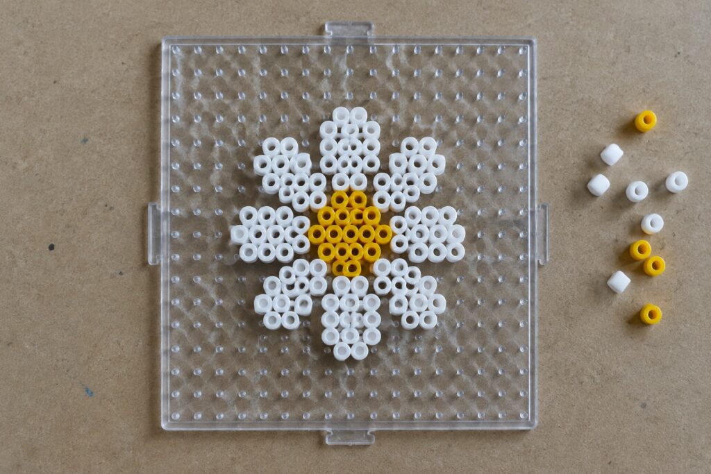 16 Daisy Perler Bead Patterns With Cheerful Petal Designs Daisy of white and yellow beads on clear pegboard with loose beads nearby.