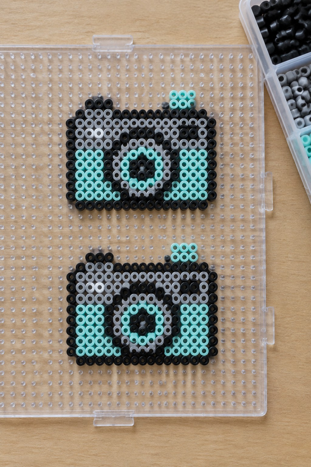 Two black, gray, white, and cyan fuse-bead pixel designs on a clear pegboard beside a bead organizer.