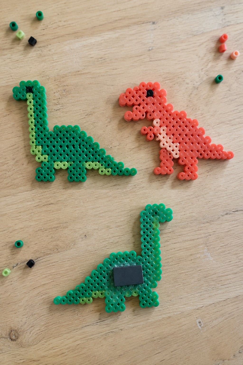 Three Perler bead dinosaurs in green and orange on wooden surface with loose beads.