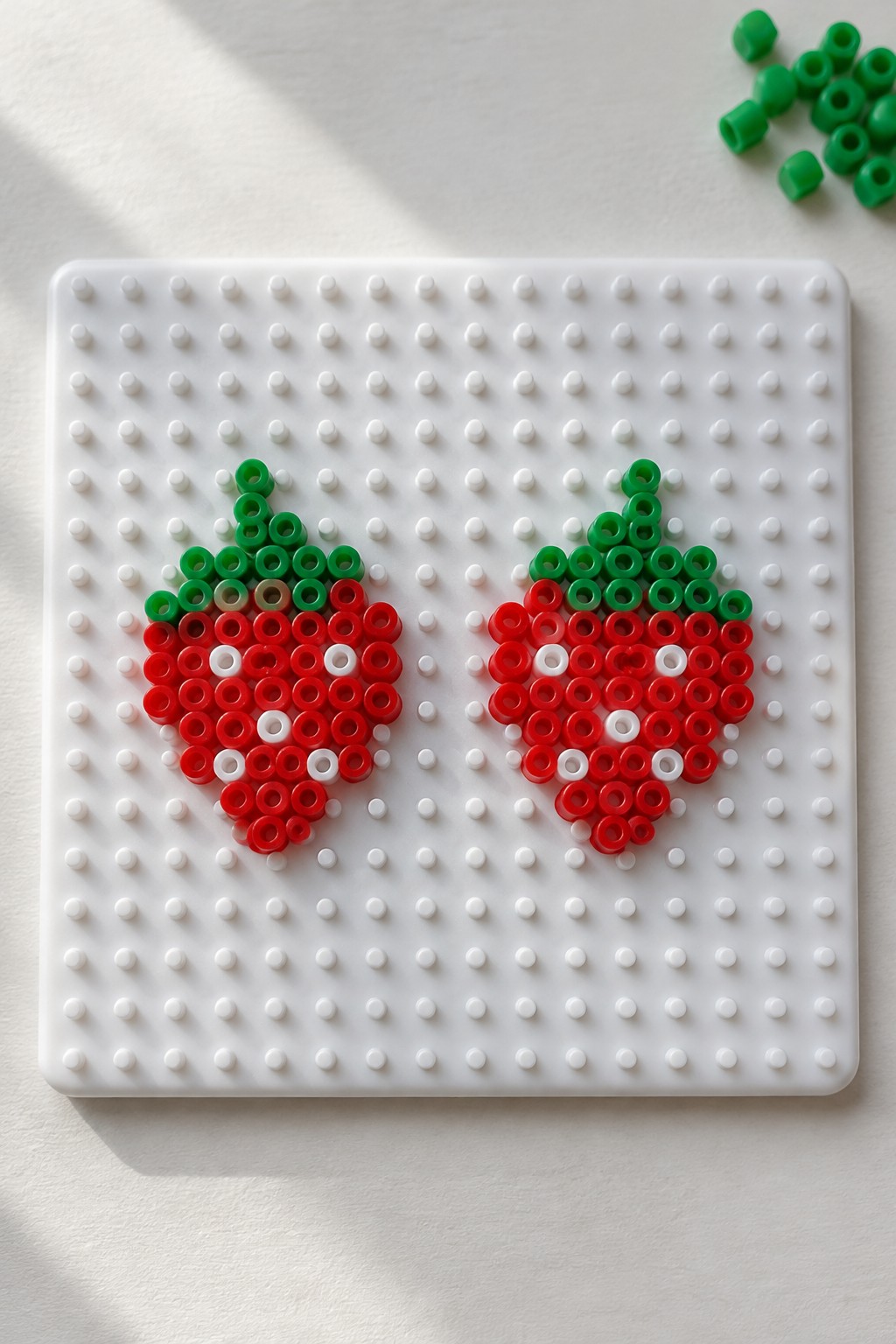 Two strawberry-shaped Perler bead patterns made with red, green, and white beads on a white pegboard.