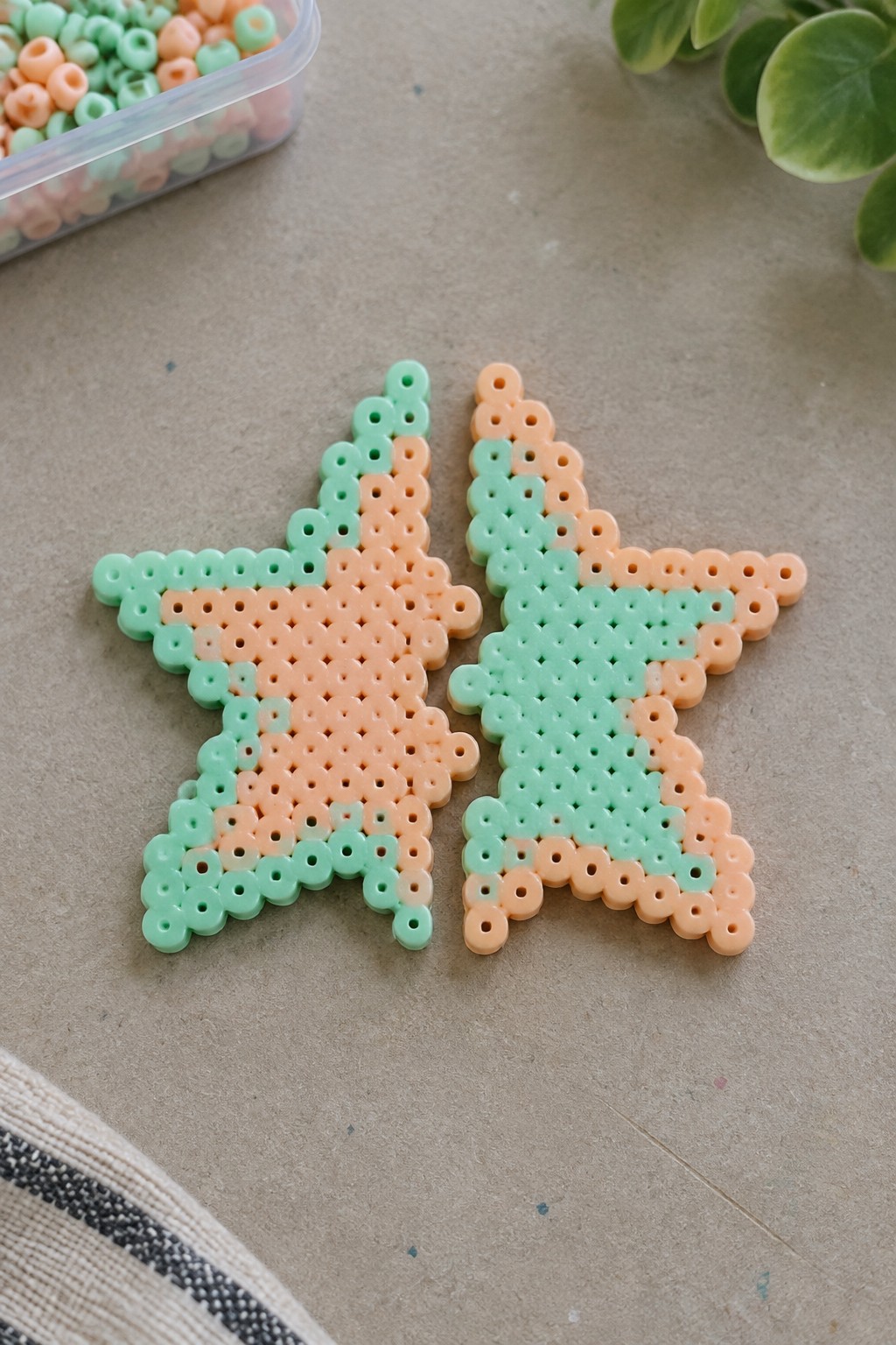Two mint green and peach perler bead stars on beige surface with beads and plant