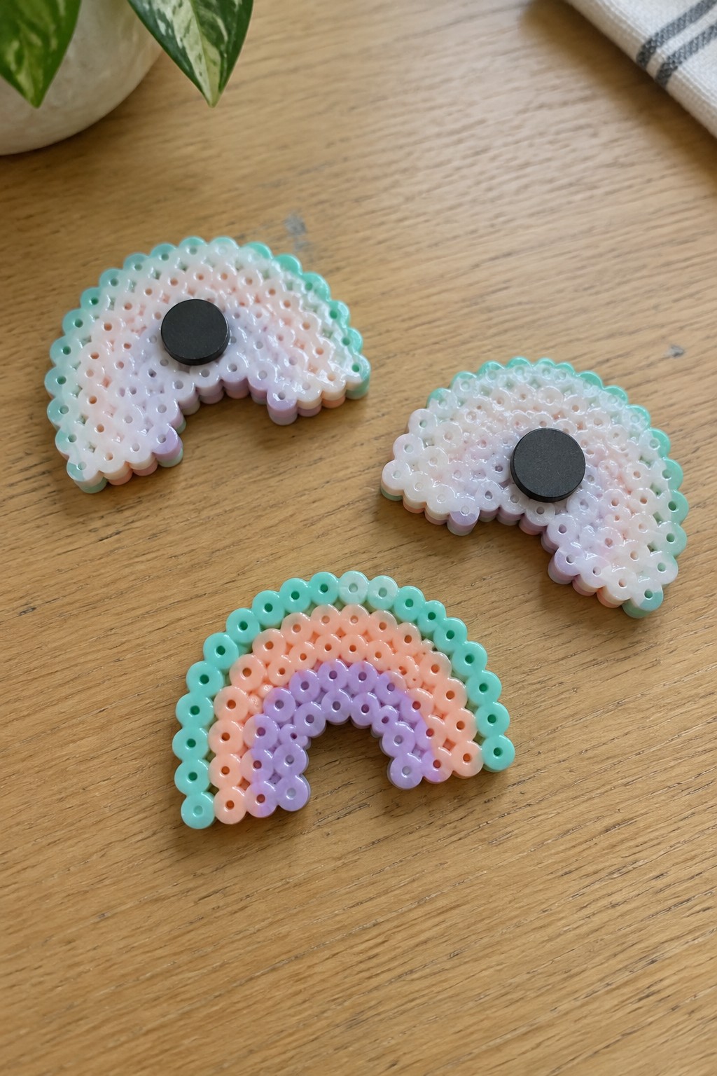 Three pastel perler bead rainbows with black magnets on wooden table