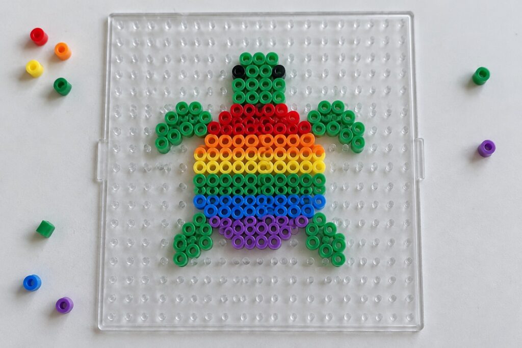 16 Rainbow Turtle Perler Bead Ideas With Bright Shell Patterns Rainbow perler bead turtle with green limbs on clear pegboard with scattered beads