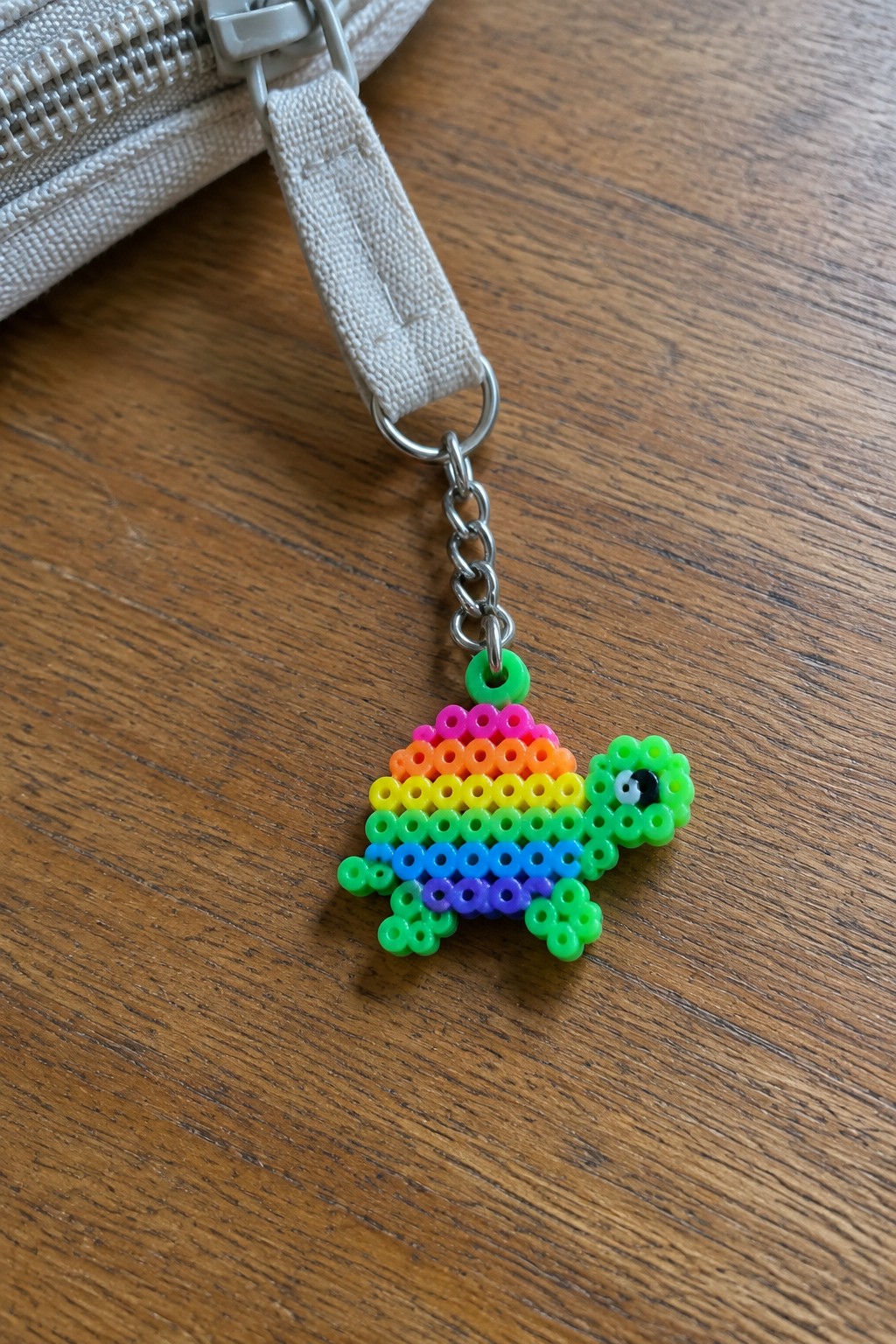 Rainbow perler bead turtle keychain dangling from white strap on wood