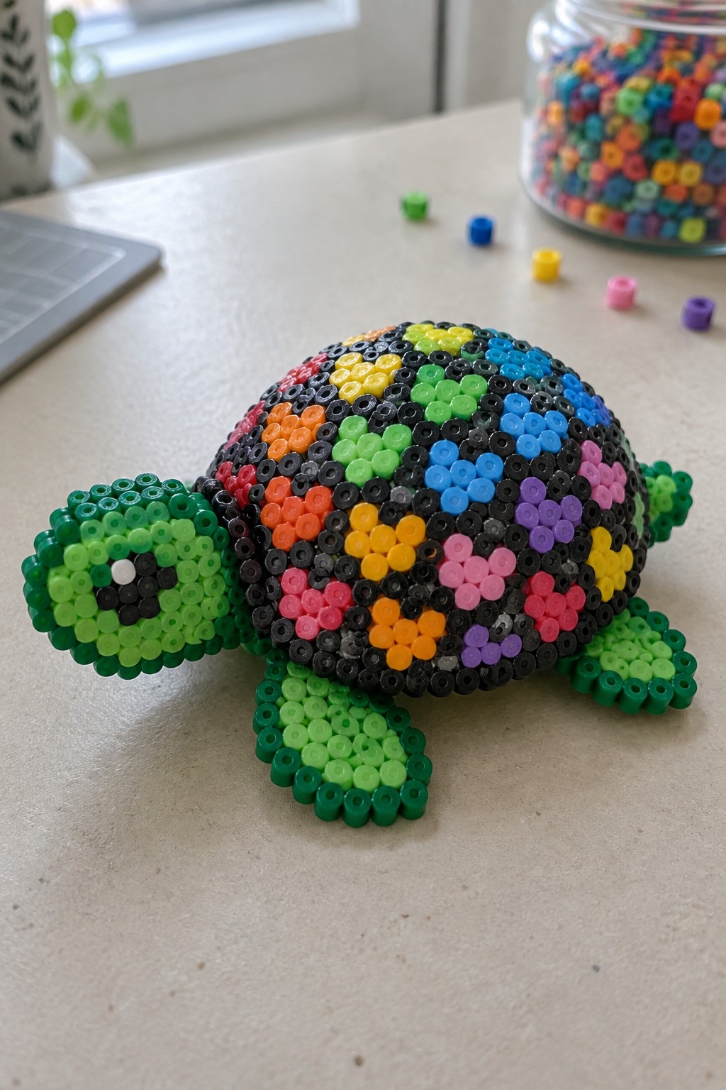 Colorful Perler bead turtle with patterned shell on light table beside bead jar.