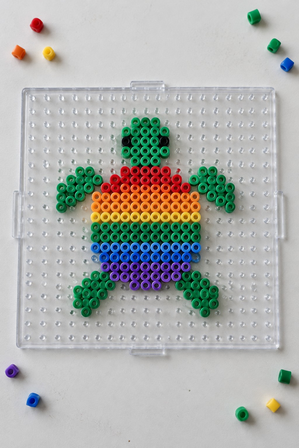 Rainbow perler bead turtle with green limbs on clear pegboard with scattered beads