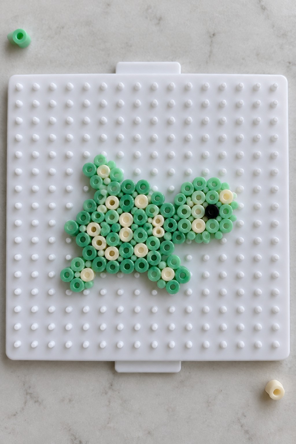 Perler bead turtle arranged on a white pegboard using multiple green shades, cream beads, and one black bead for the eye.