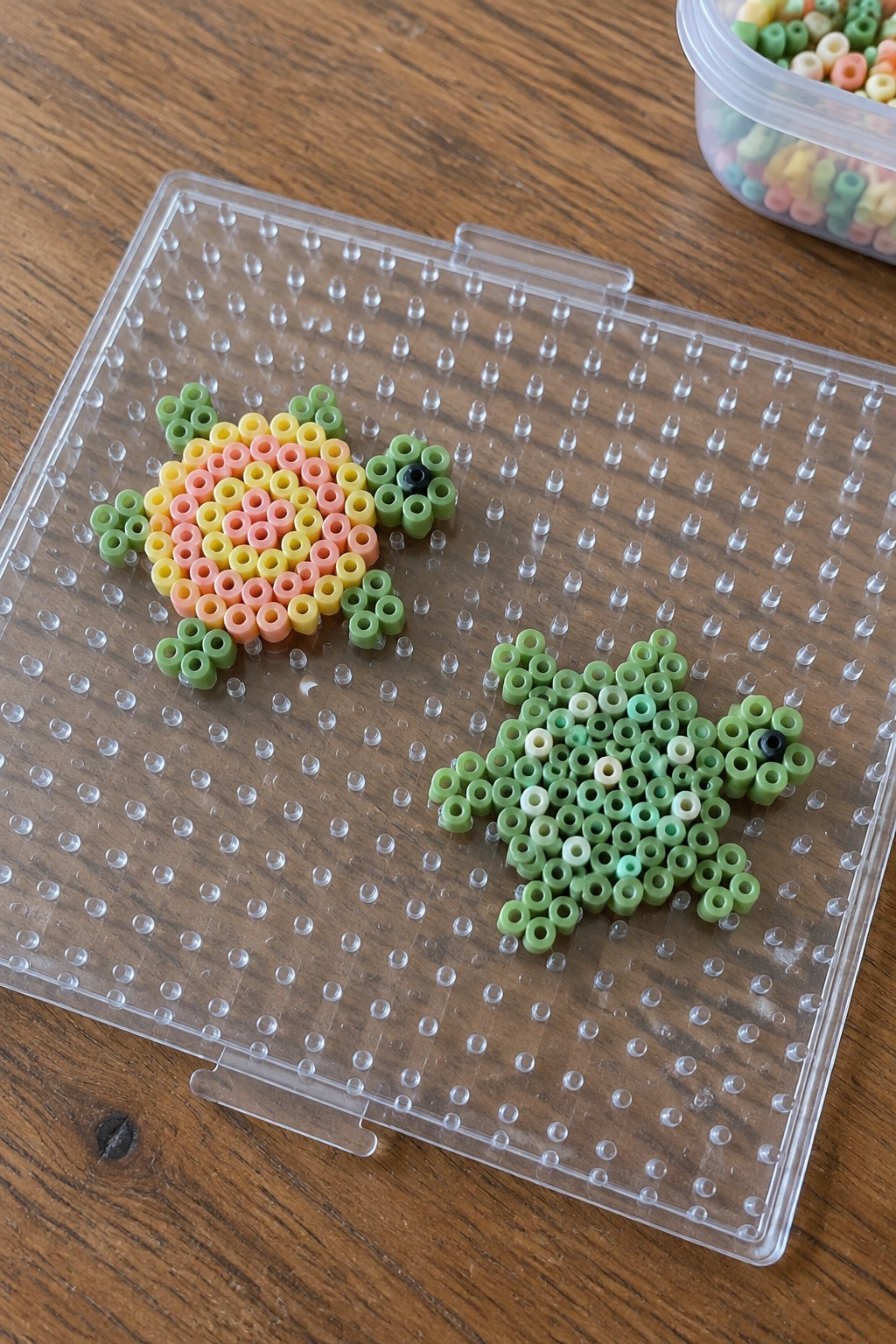 Two perler bead turtles, rainbow and green, on clear pegboard over wood