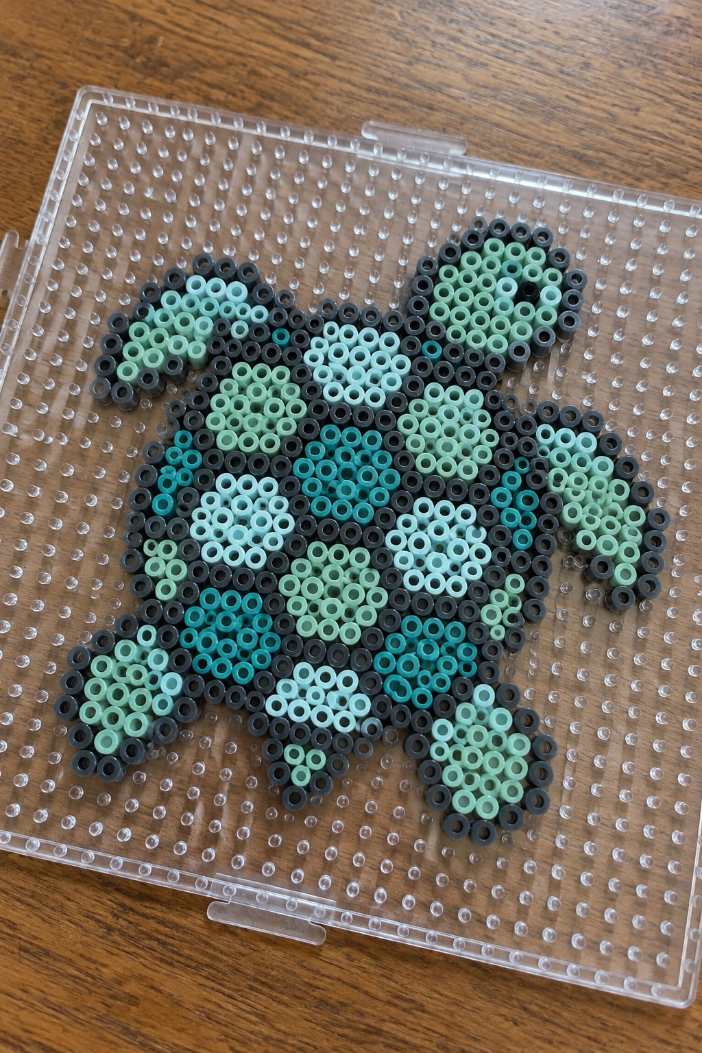 Pixelated creature made of green, teal, and black perler beads on pegboard
