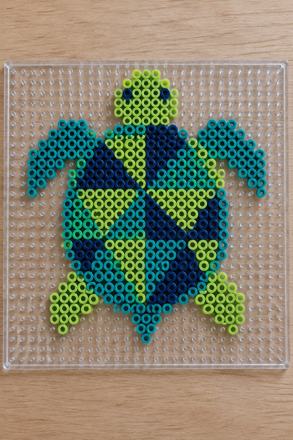 Colorful perler bead turtle in green and blue on clear pegboard
