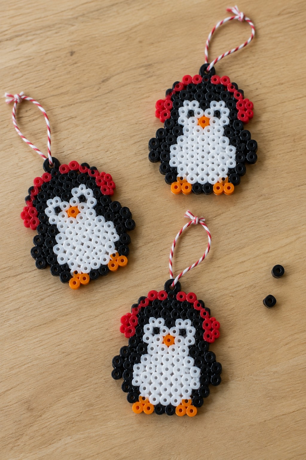 Three perler bead penguin ornaments in black, white, red, orange with striped strings on wood.
