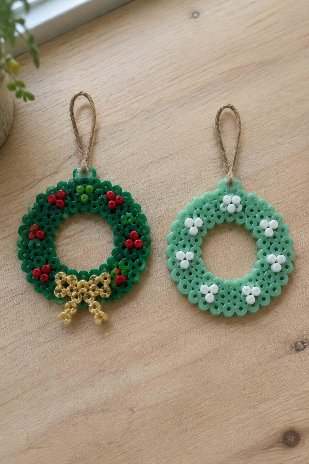 Two fused Perler bead Christmas wreath ornaments on a wooden surface, one dark green with red beads and gold bow, the other light green with white accents, both with twine loops.