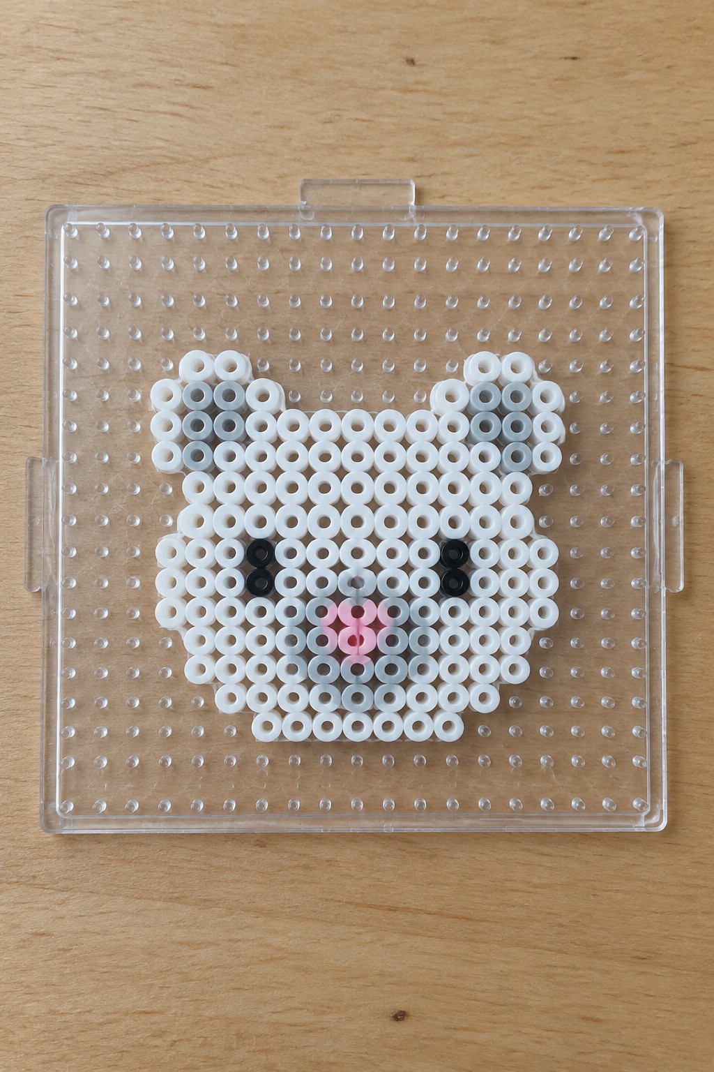 Perler beads forming white cat face with black eyes and pink nose on clear pegboard