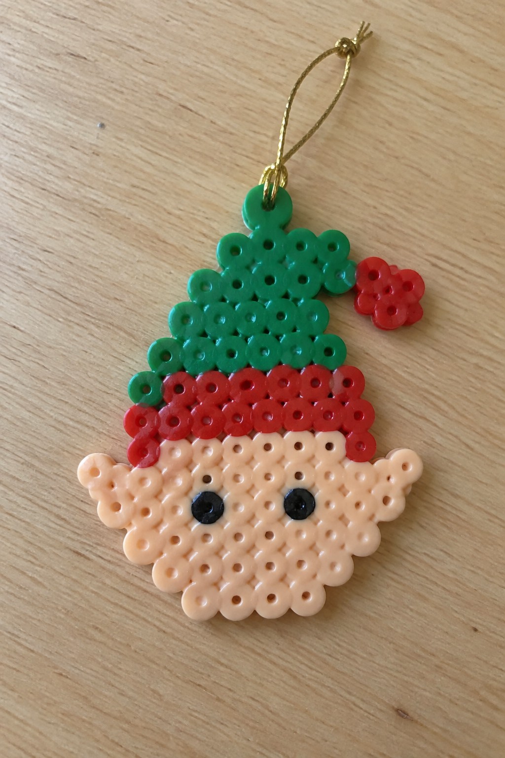 Perler bead elf ornament with green hat, red brim, pom-pom, and black eyes on wood