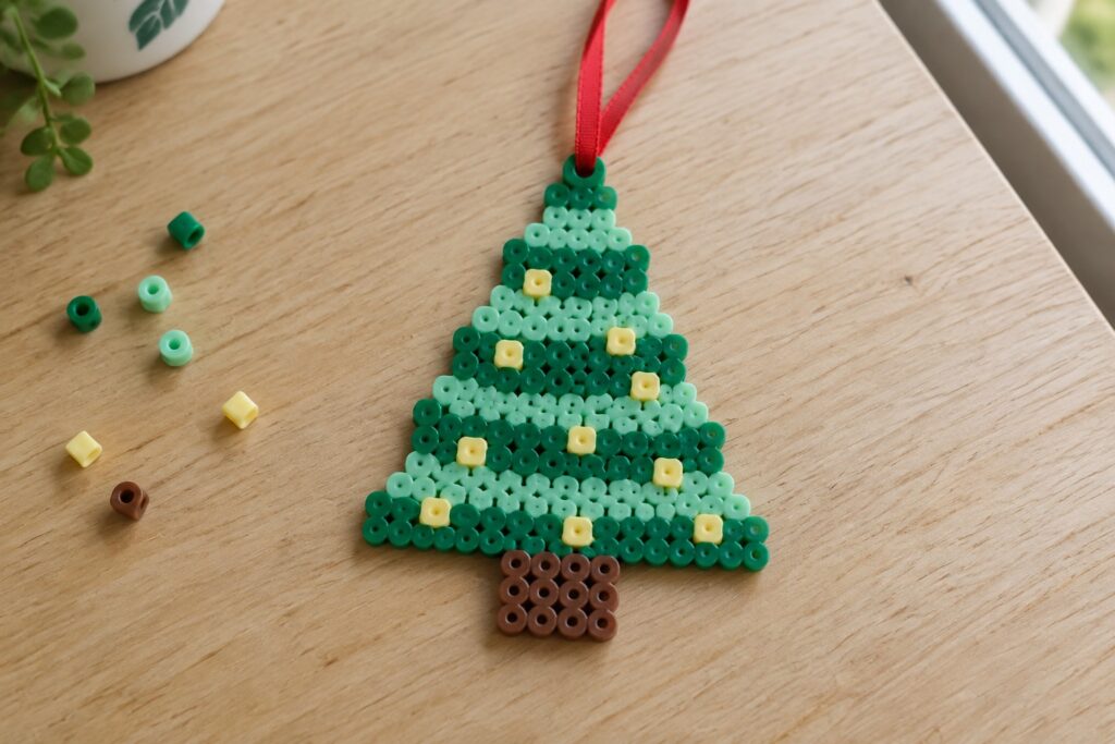 17 Cute Christmas Perler Bead Ideas for Festive Ornaments Green perler bead Christmas tree ornament with yellow lights, brown trunk, red ribbon on wood.