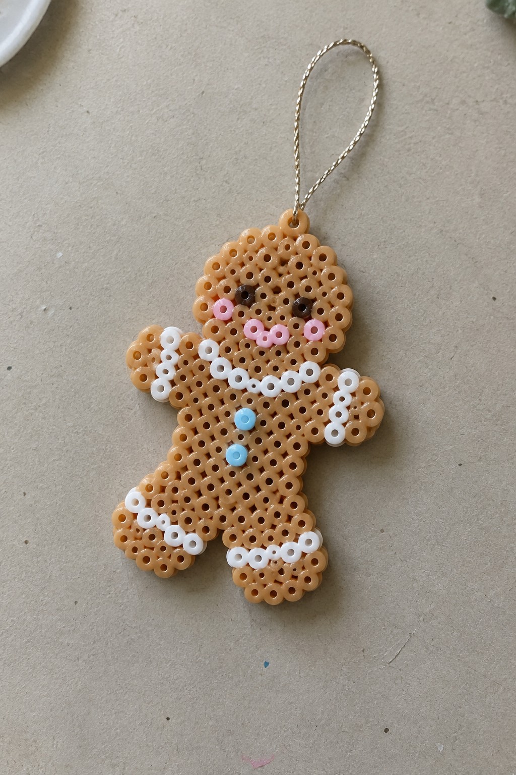 Perler bead gingerbread man ornament with white icing, pink cheeks, blue buttons, gold loop