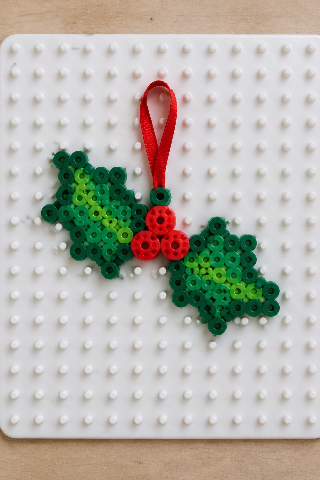 Perler bead holly leaves with red berries and ribbon on pegboard