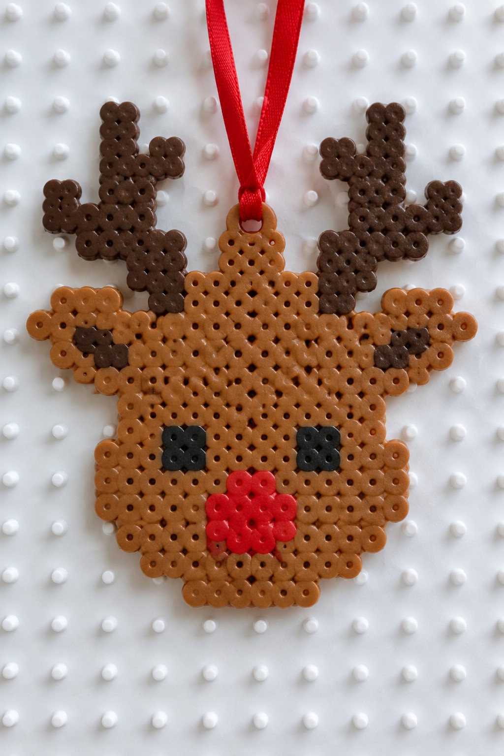 Brown perler bead reindeer ornament with antlers, black eyes, red nose, and ribbon on pegboard.