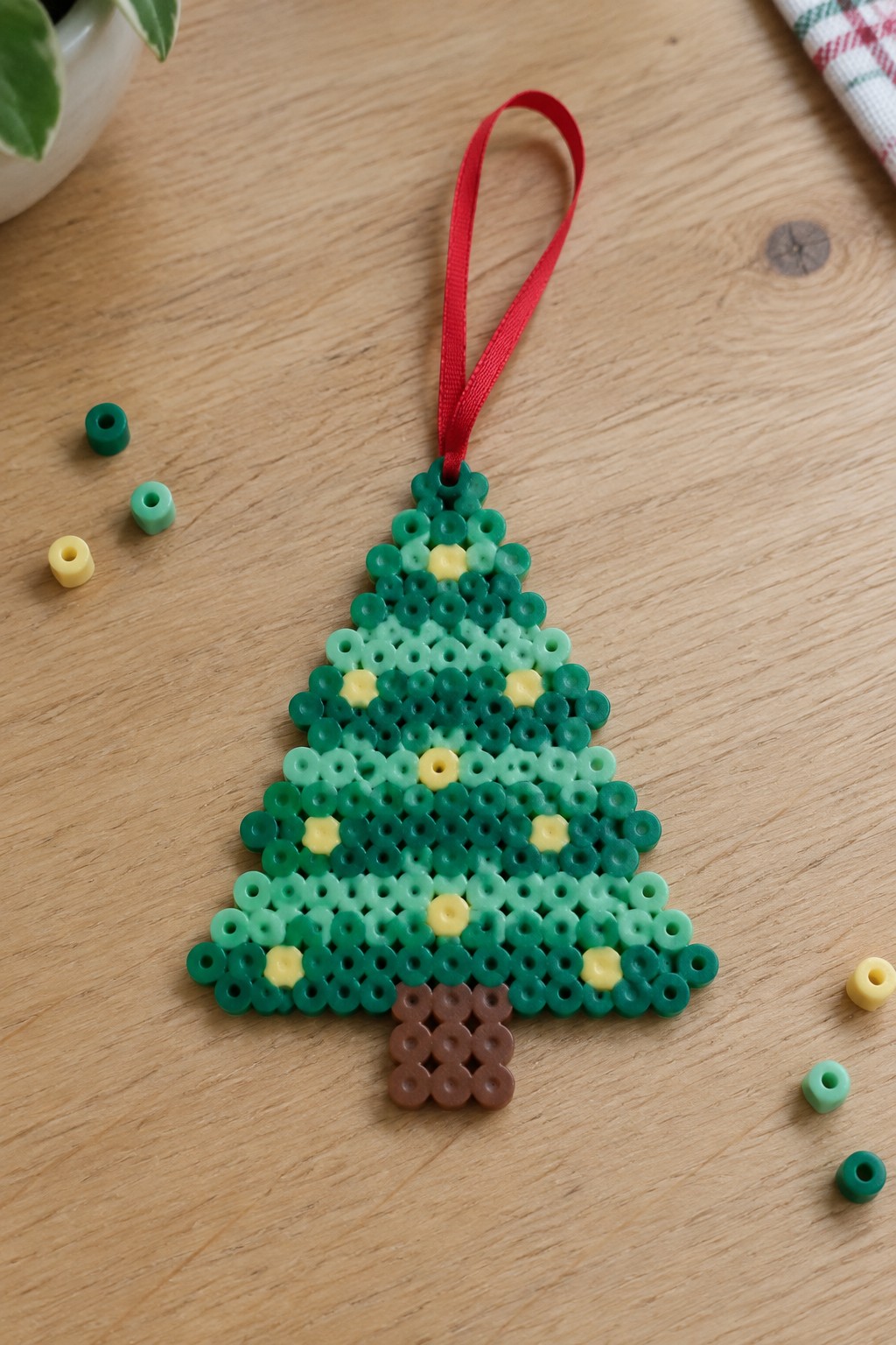 Green perler bead Christmas tree ornament with yellow lights, brown trunk, red ribbon on wood.