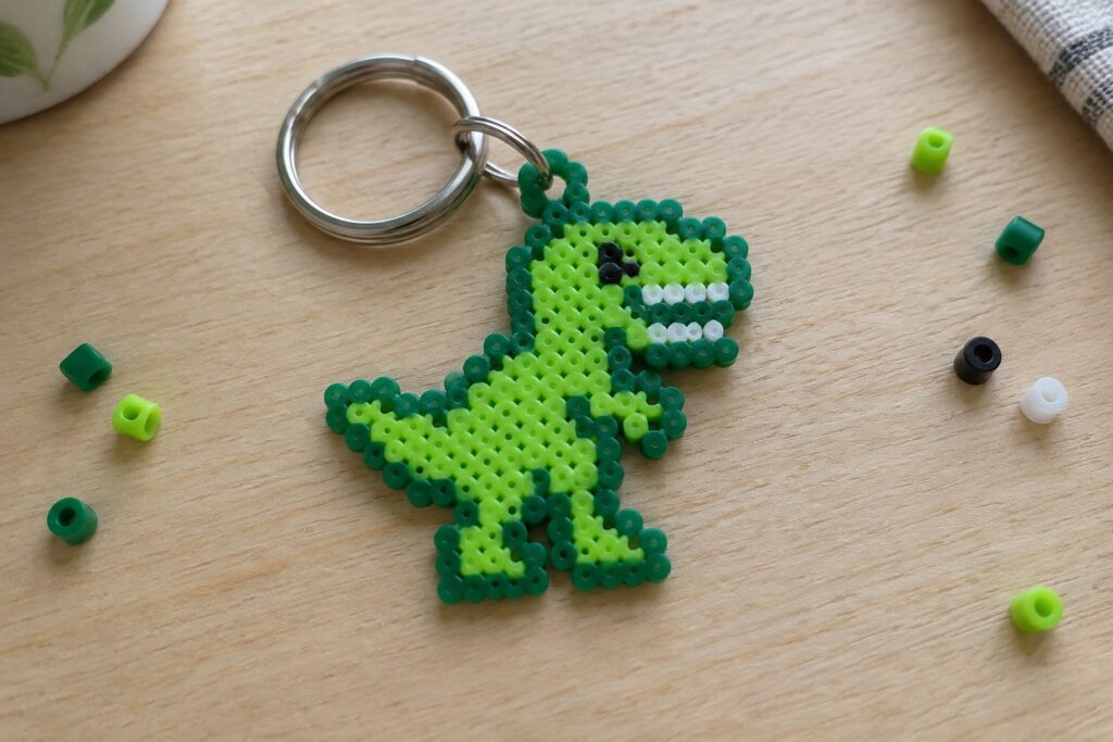 Green perler bead dinosaur keychain on wooden table with scattered beads