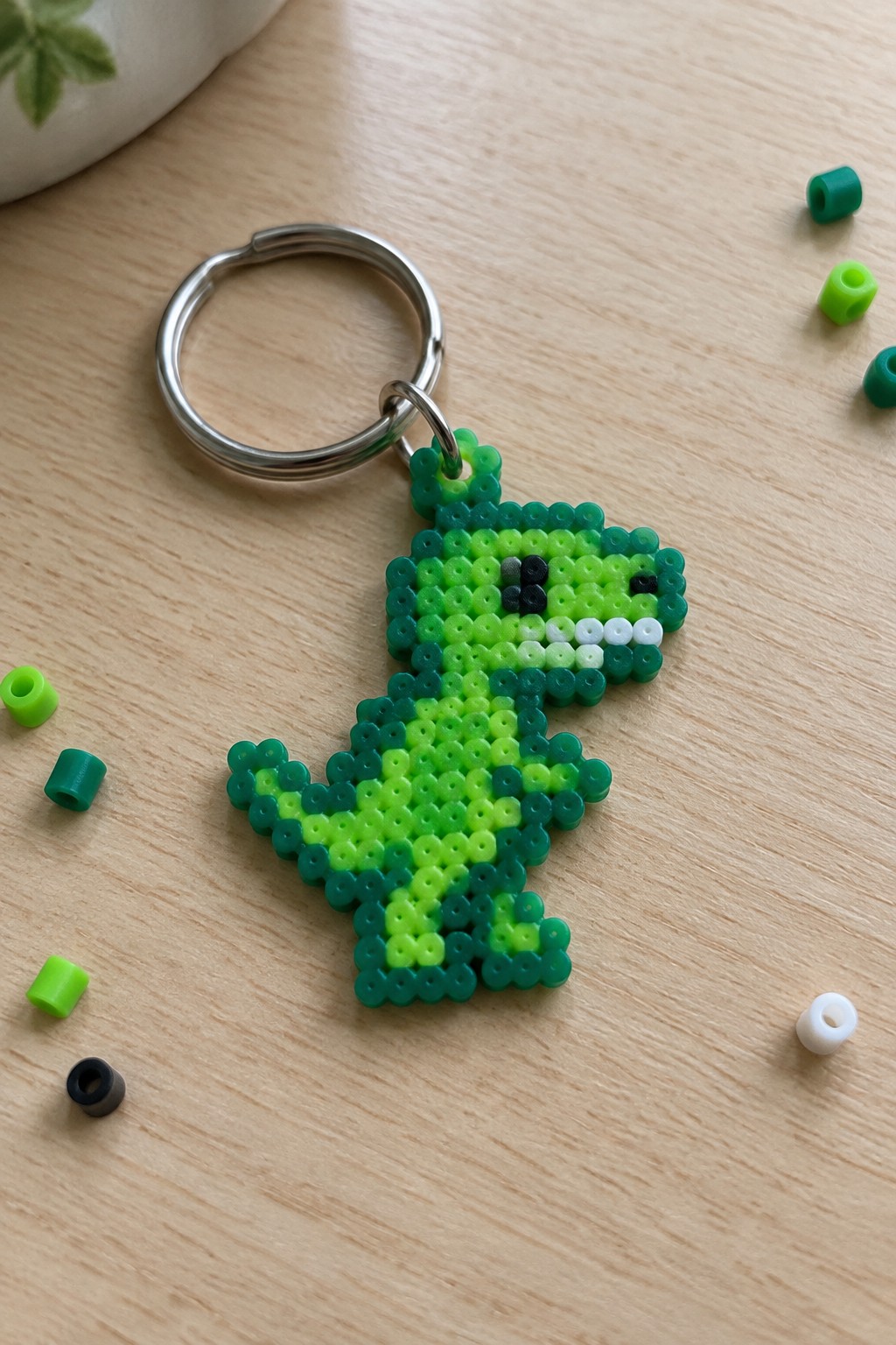 Green perler bead dinosaur keychain on wooden table with scattered beads