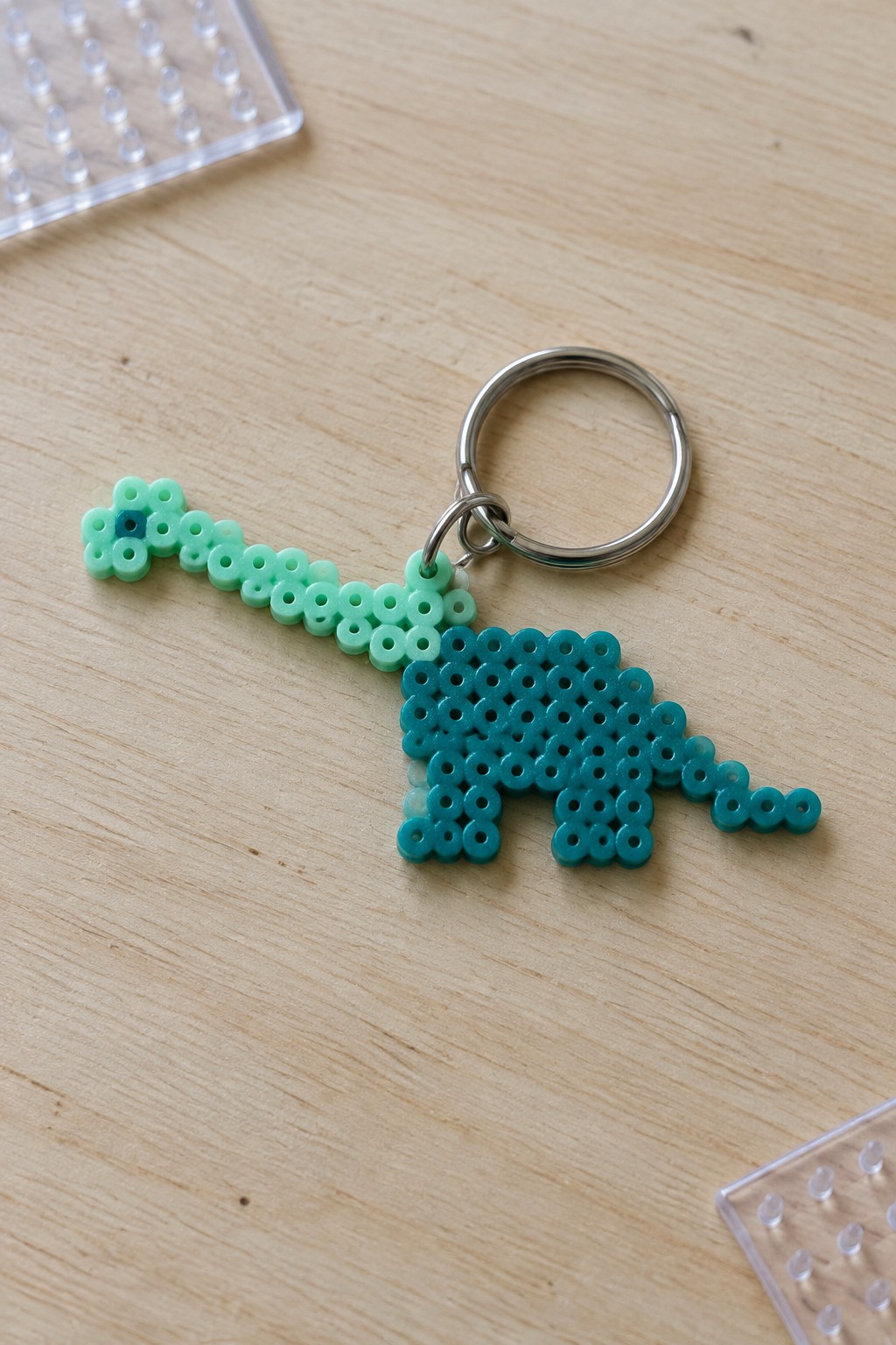 Mint and teal perler bead dinosaur keychain on silver ring atop wood