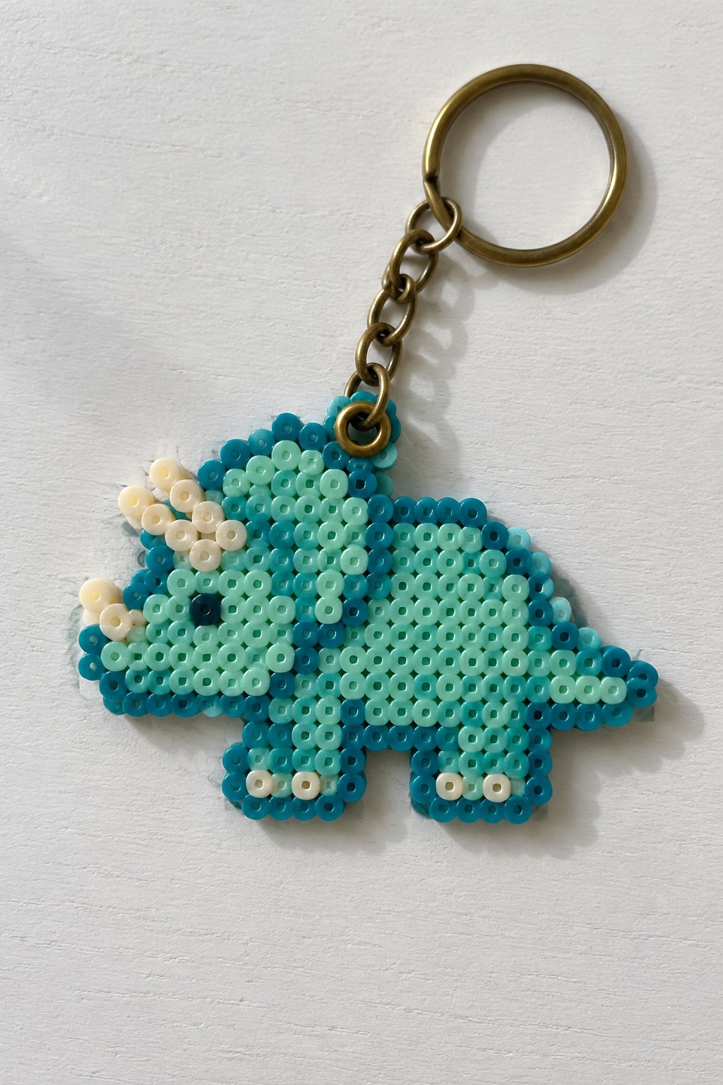 Fused Perler bead triceratops keychain in blue and teal beads attached to a brass keyring.