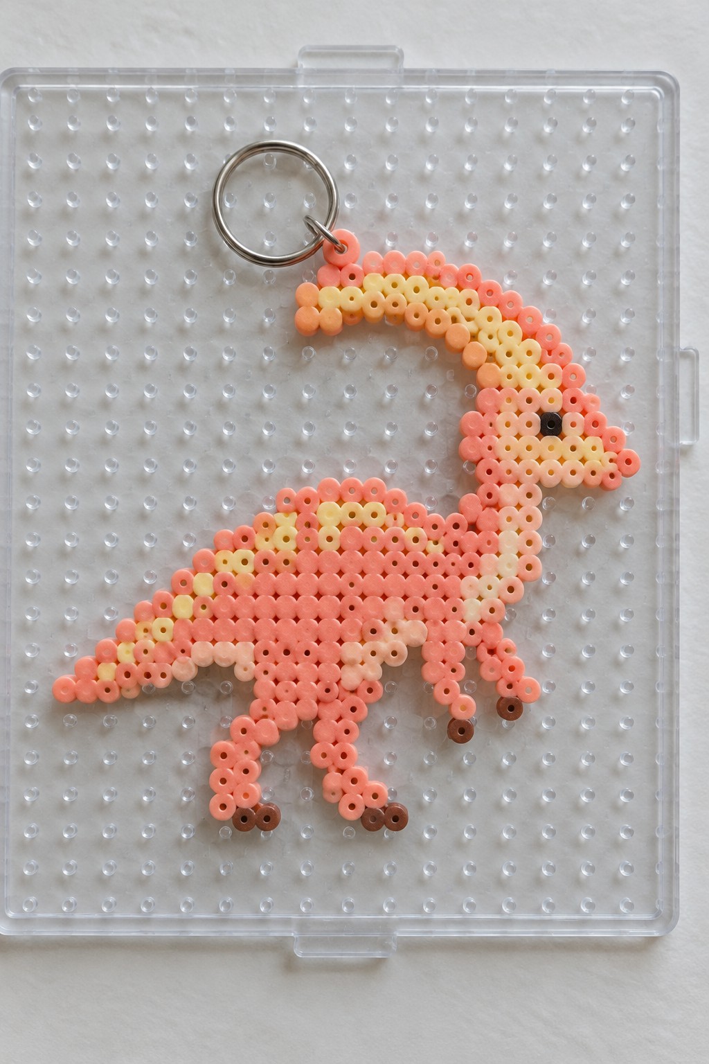 Pink perler bead dinosaur keychain with yellow crest on clear pegboard.