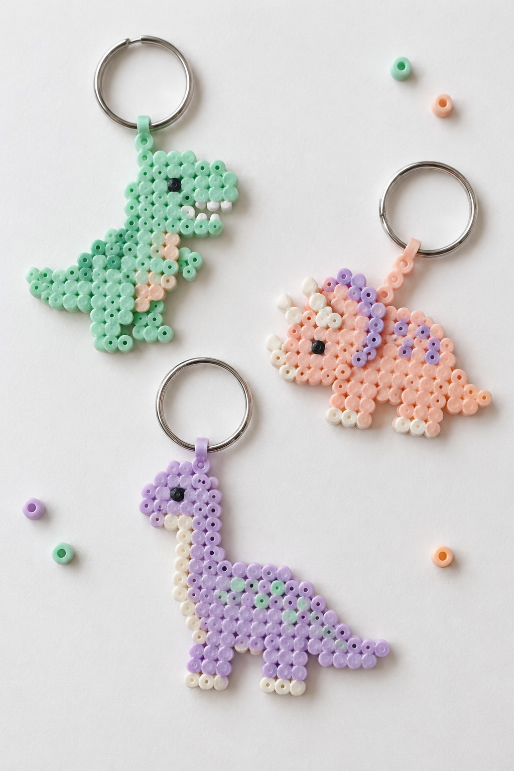 Three fused Perler bead dinosaur keychains in mint green, peach with purple, and lavender with white.
