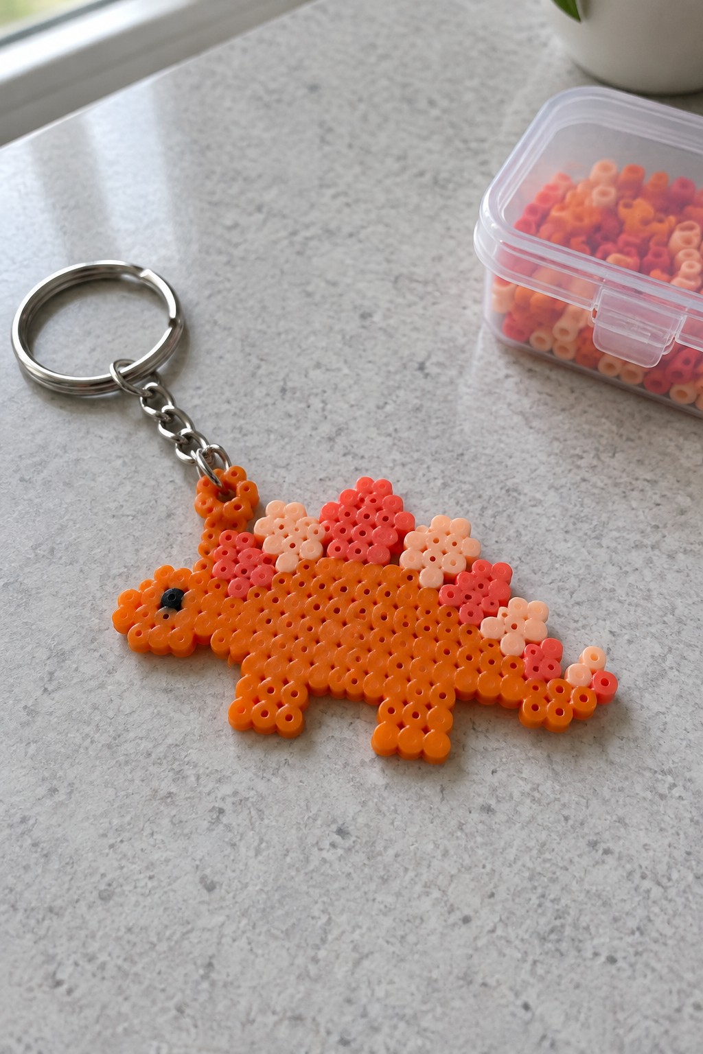 Orange perler bead stegosaurus keychain on gray countertop beside bead container