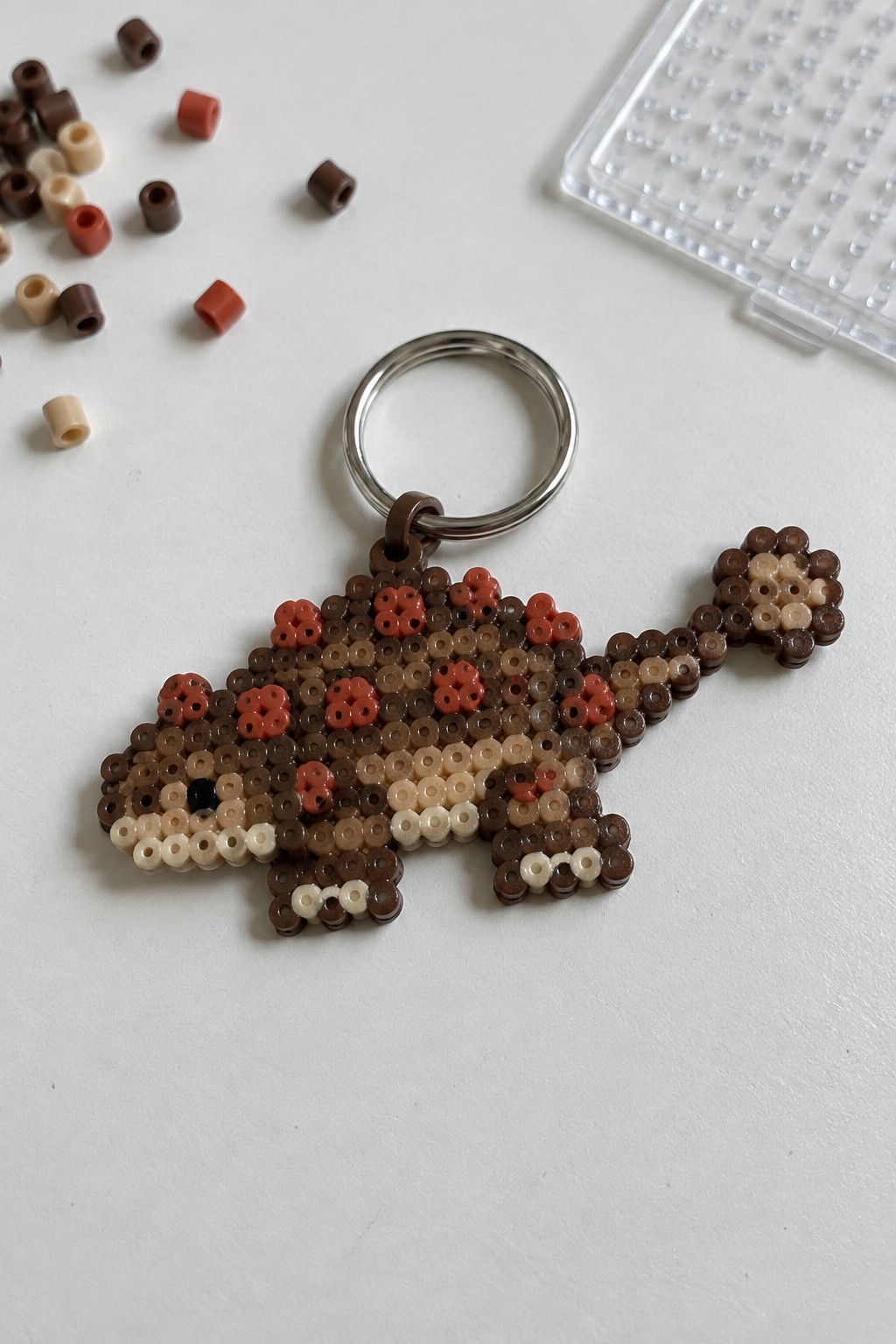 Perler bead animal keychain in brown tones with metal ring amid loose beads and pegboard.