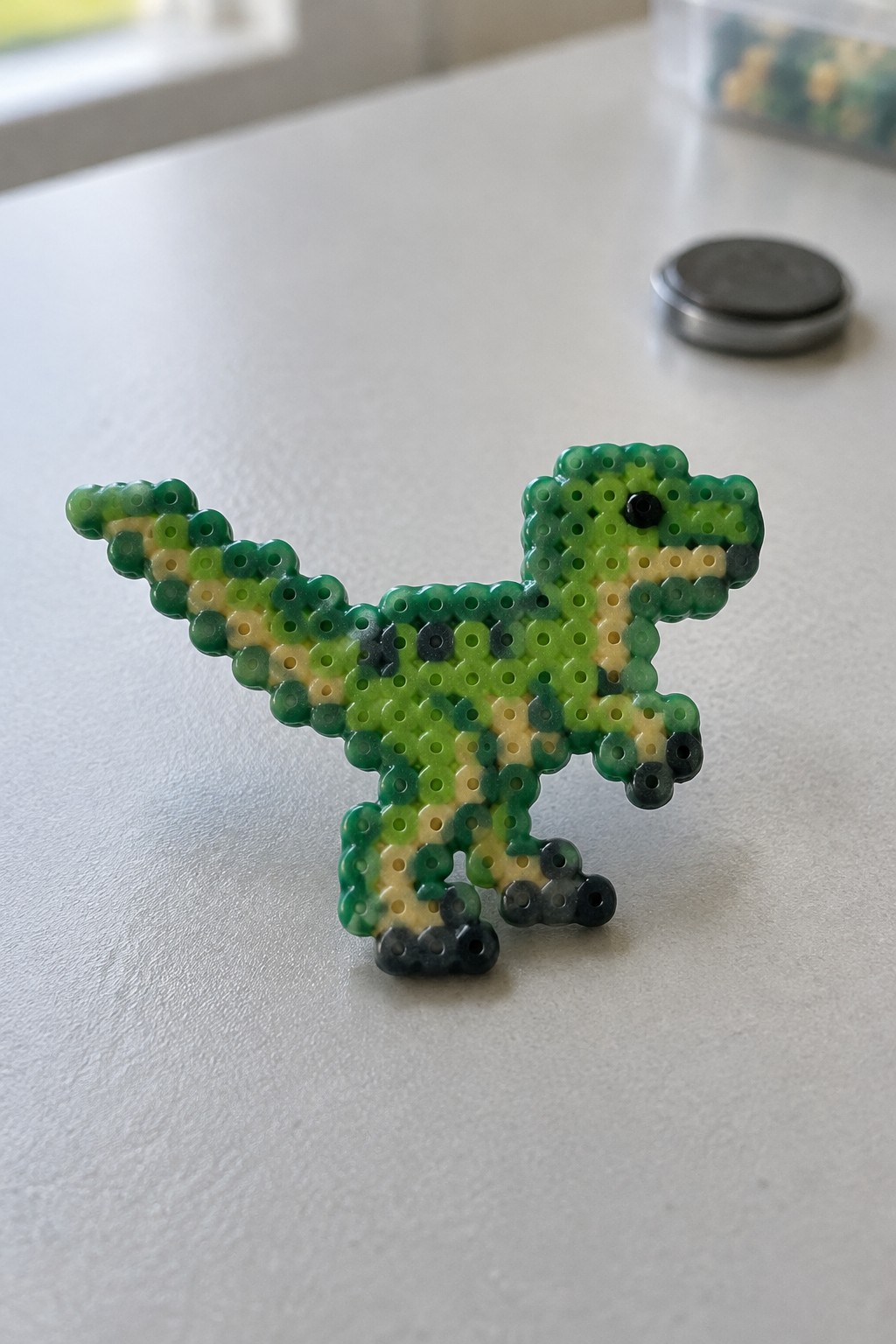 Green and yellow perler bead dinosaur on white surface beside coin
