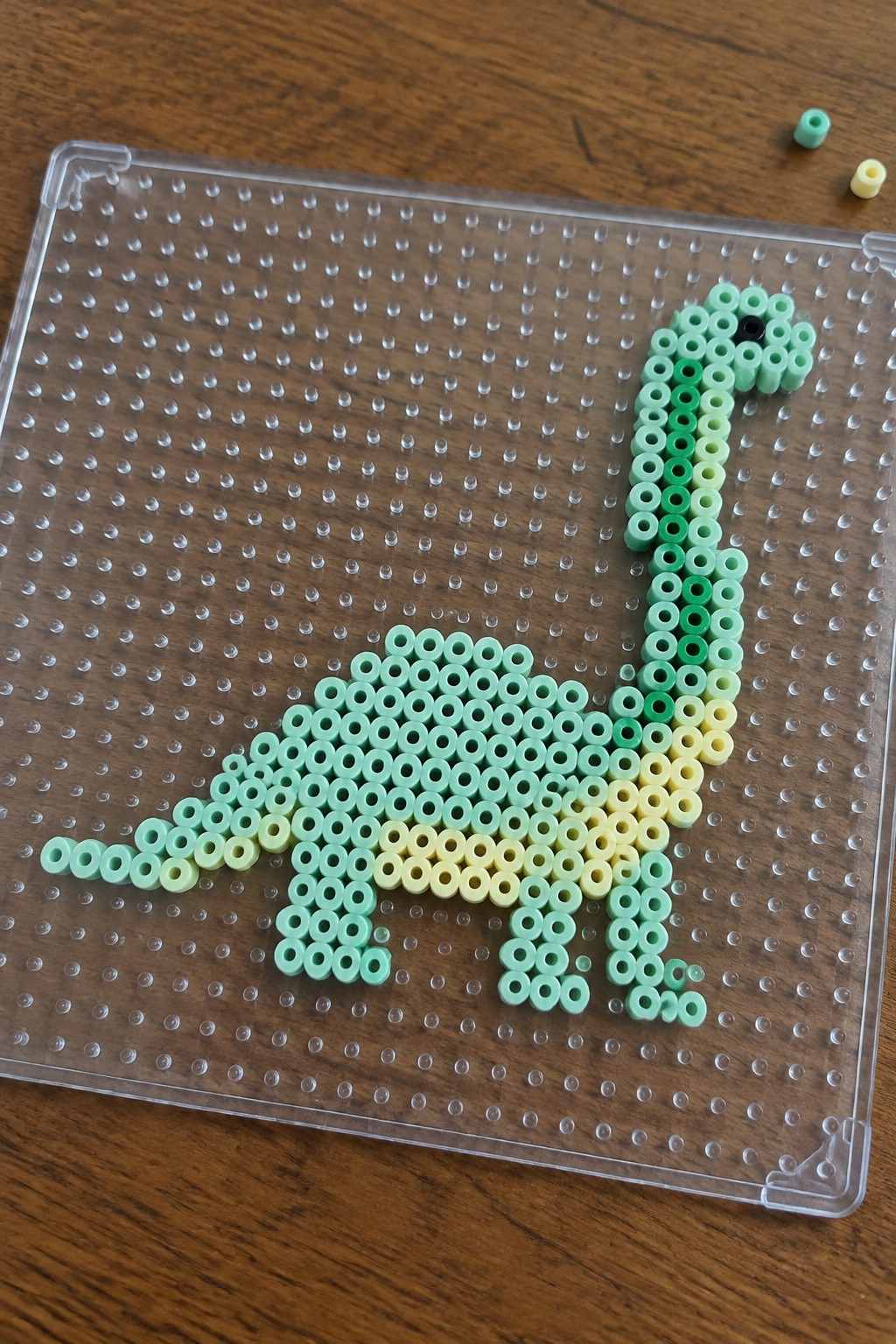 Green-yellow perler bead dinosaur on clear pegboard atop wooden table