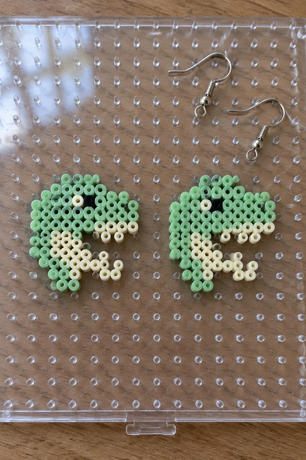 Two green perler bead frogs beside silver earring hooks on clear pegboard