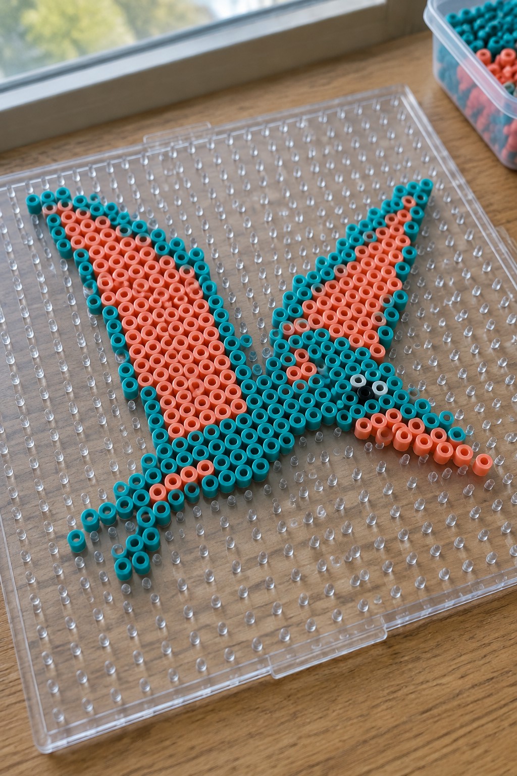 Orange and teal perler beads forming a bat on a clear pegboard.