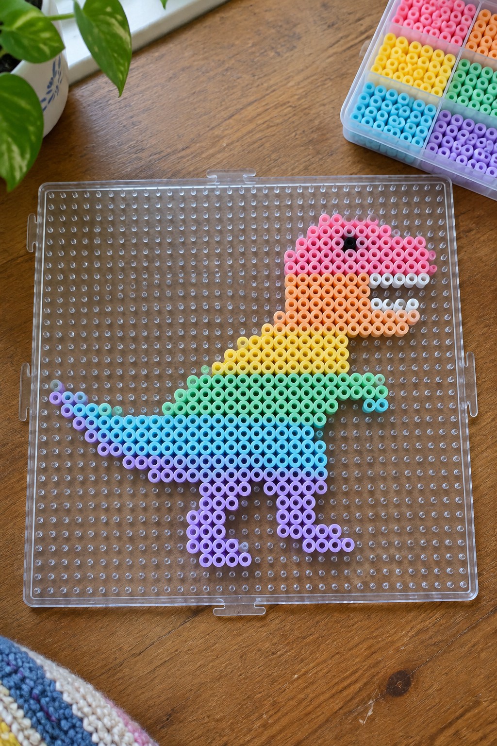 Rainbow perler bead dinosaur on clear pegboard with colorful bead organizer nearby