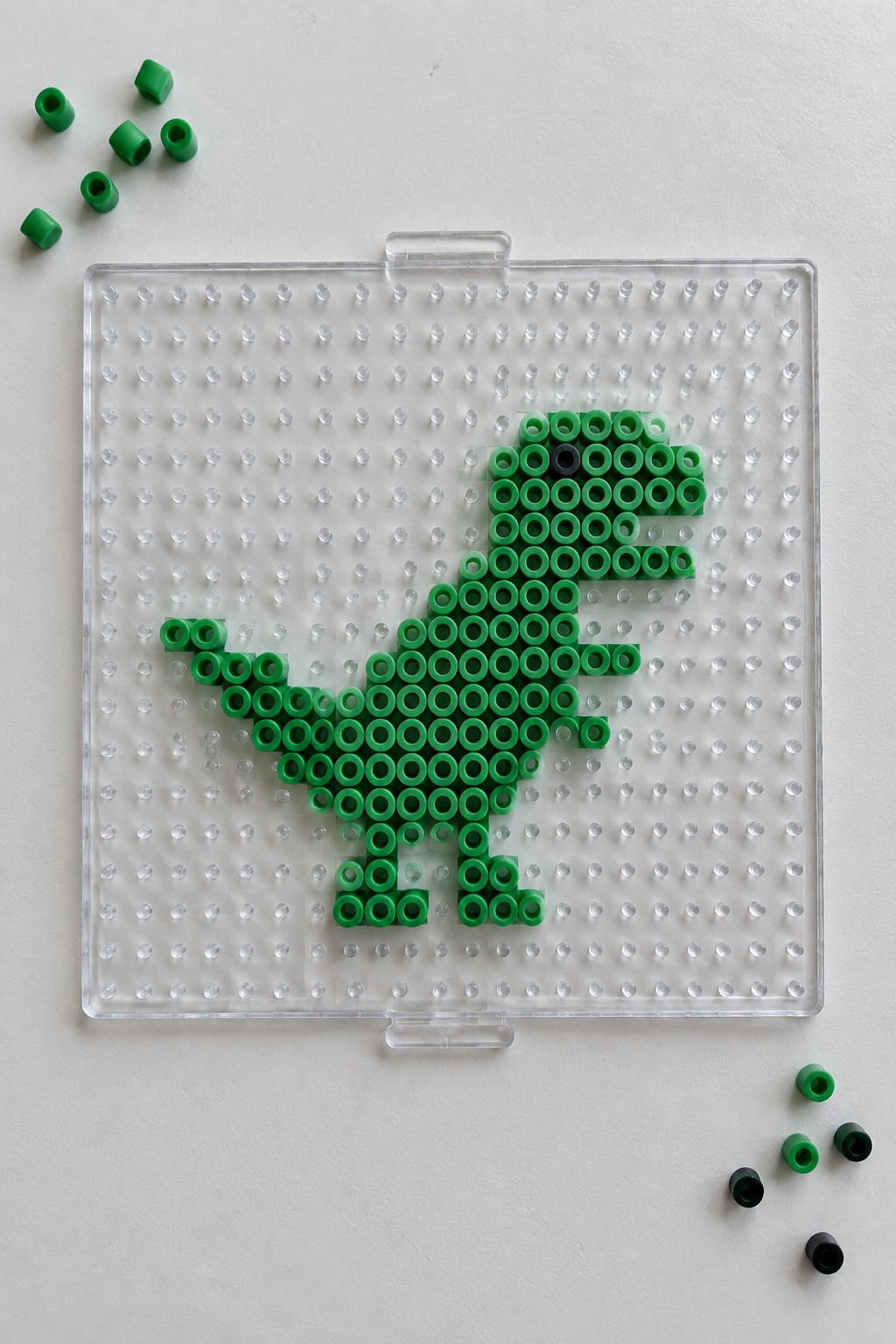 Green perler bead T-Rex on transparent pegboard with loose beads nearby