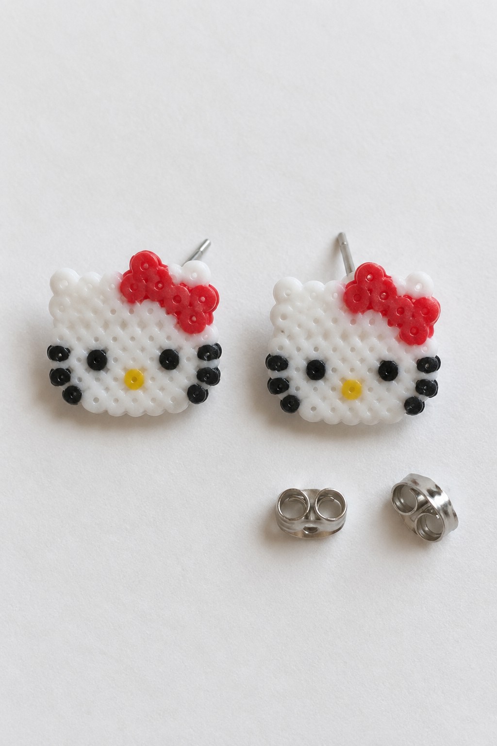 Two small Hello Kitty faces made from white, red, black, and yellow Perler beads, each fitted with a metal earring post, shown with backs.