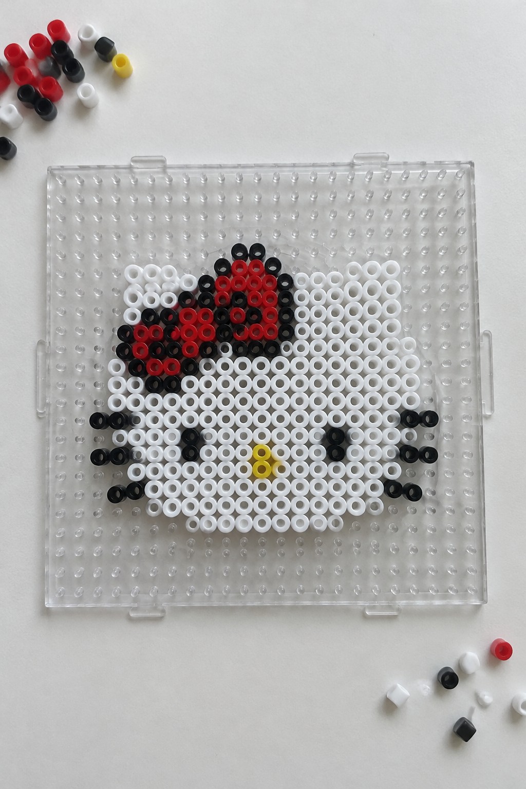 Perler beads forming Hello Kitty face on clear pegboard with scattered loose beads nearby.
