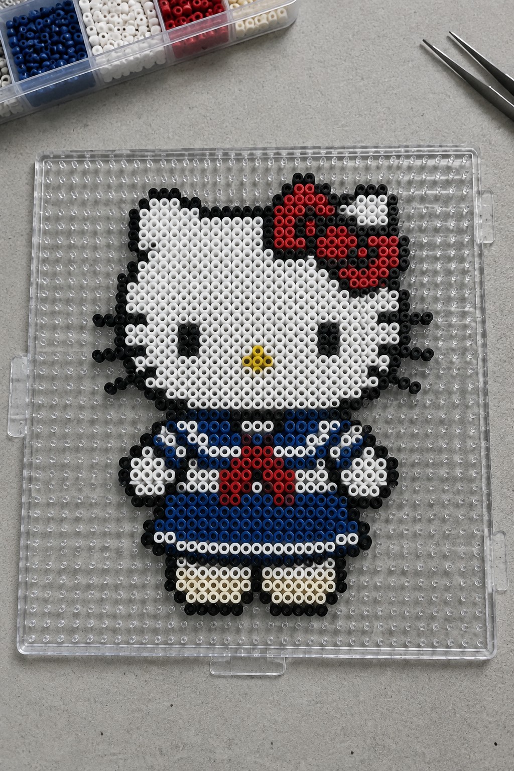 Hello Kitty perler bead art on clear pegboard with bead trays and tweezers nearby