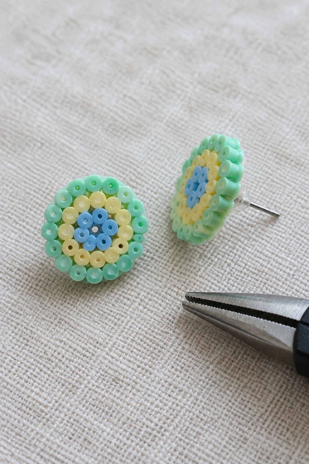 Two pastel beaded earrings and metal pliers on white textured fabric