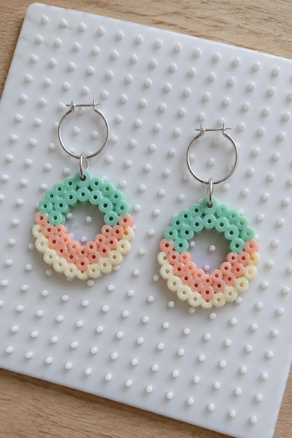 Two Perler bead earrings with mint green, peach, and cream circular ring designs on silver hoops resting on a white pegboard.