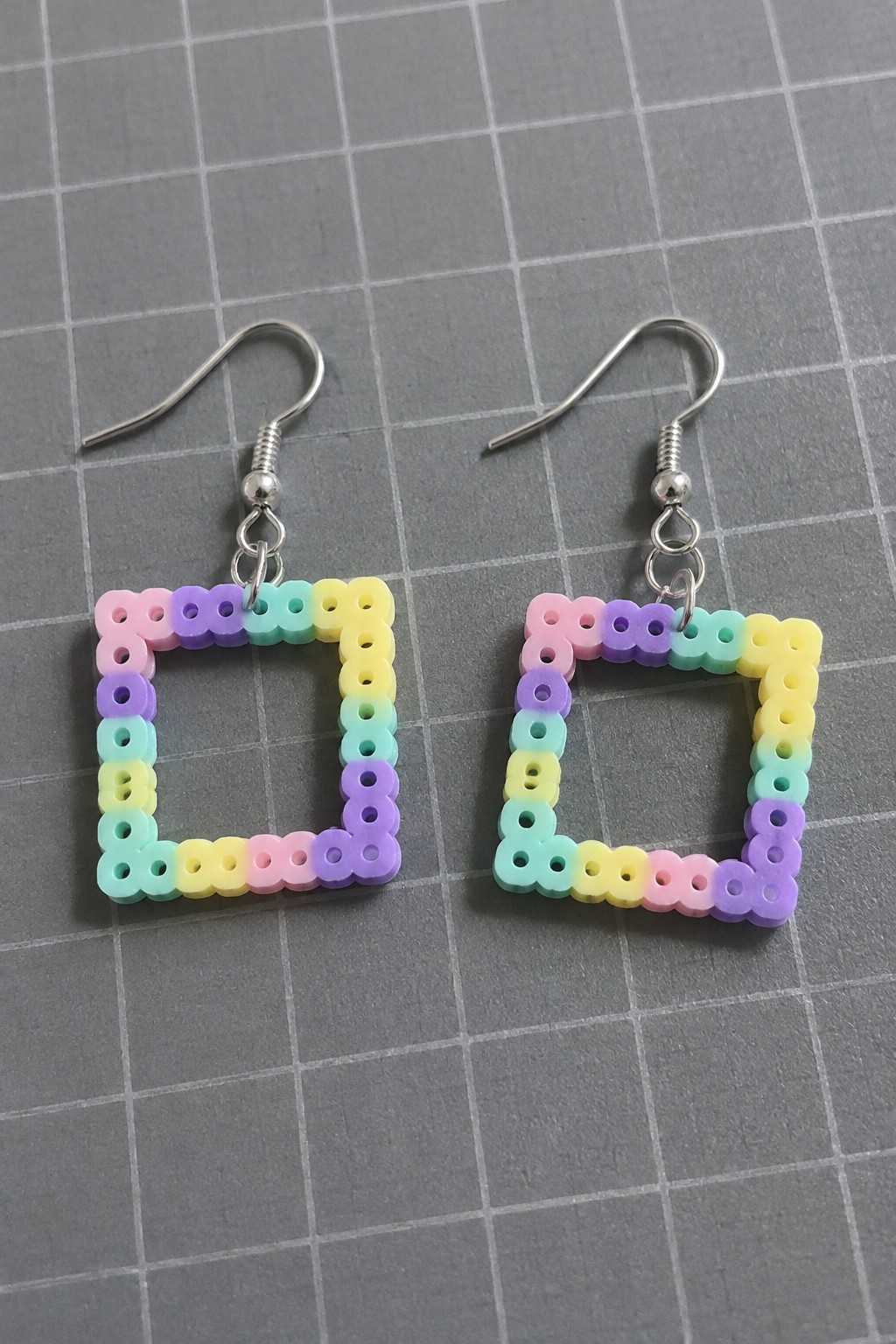 Pair of square rainbow perler bead earrings with silver hooks on gray grid.