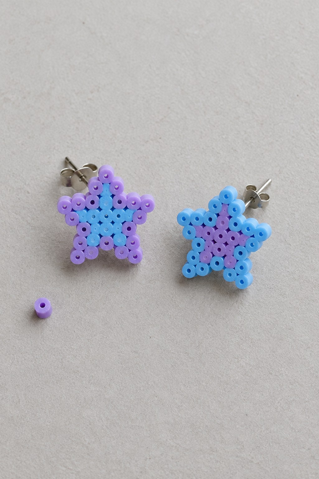 Two perler bead star earrings in purple and blue beside a loose bead